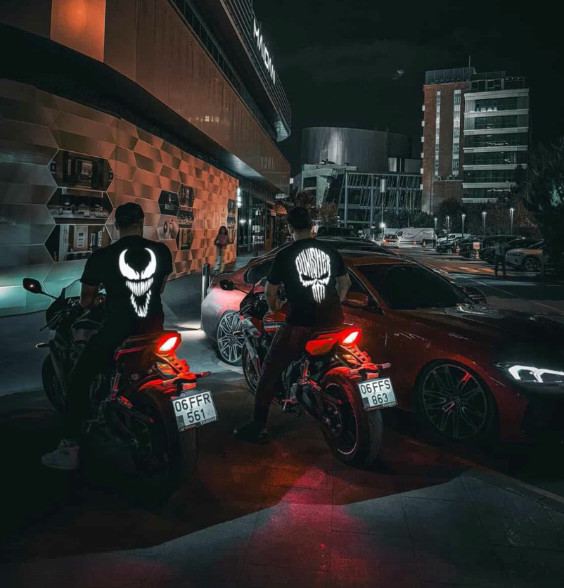 Gece iki motosiklet sürücüsünün sırtında parlayan Punisher ve Venom reflektör baskılı tişörtler