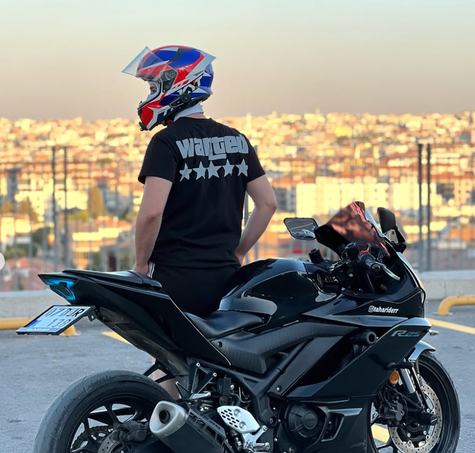Şehir manzarası önünde motosiklet üzerinde Wanted baskılı siyah tişört giyen sürücü