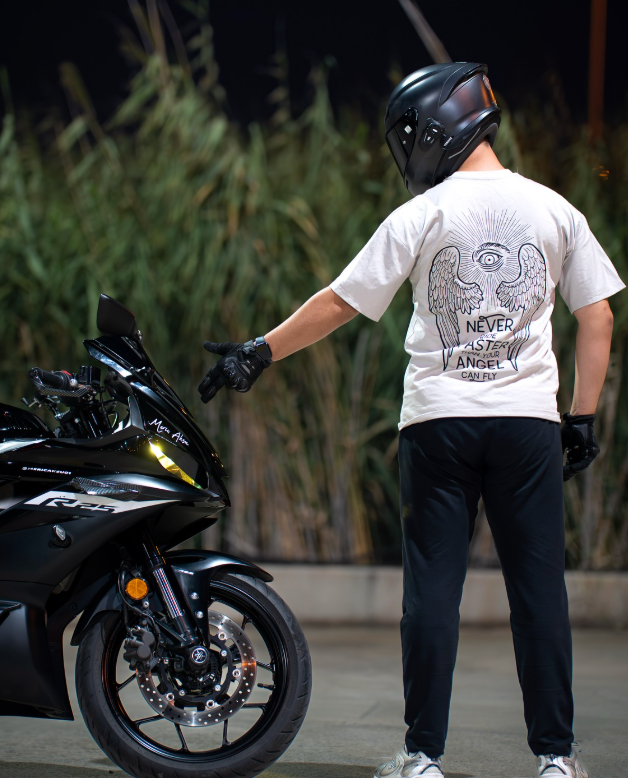 Motosikletin yanında Angel Can Fly baskılı beyaz motorcu tişört giyen kasklı sürücü